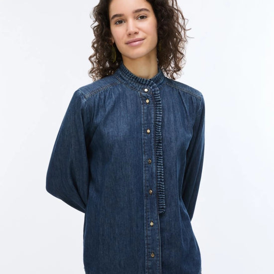 A woman with curly brown hair wearing a dark blue denim long-sleeve button-up shirt with a gathered, pleated front and collar, photographed against a plain white studio background.