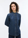 A woman with curly brown hair wearing a dark blue denim long-sleeve button-up shirt with a gathered, pleated front and collar, photographed against a plain white studio background.