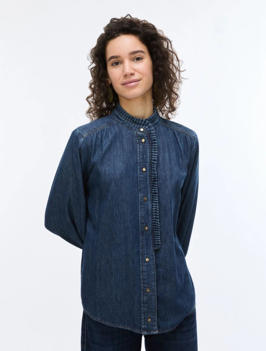 A woman with curly brown hair wearing a dark blue denim long-sleeve button-up shirt with a gathered, pleated front and collar, photographed against a plain white studio background.