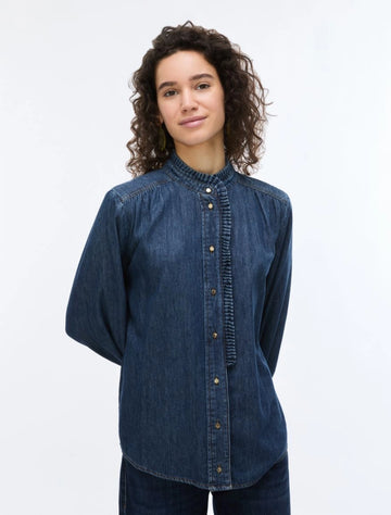 A woman with curly brown hair wearing a dark blue denim long-sleeve button-up shirt with a gathered, pleated front and collar, photographed against a plain white studio background.