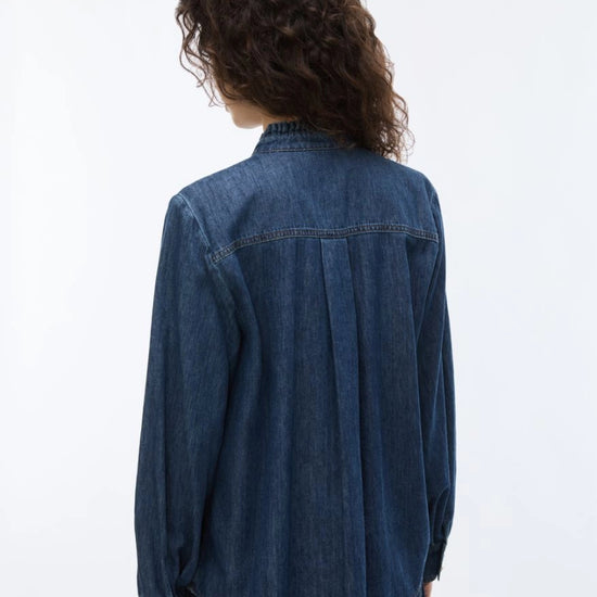 A person shown from the back wearing a dark blue denim long-sleeve button-up shirt and dark jeans against a plain white studio background.