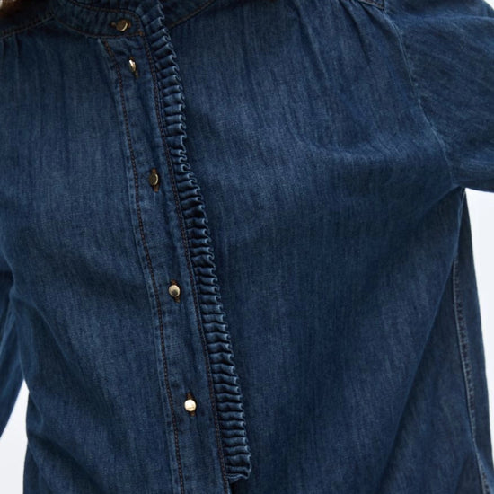 A model wearing a dark blue denim button-up shirt, matching wide-leg jeans, a brown belt, and holding a black leather jacket against a plain white studio background CLOSE UP