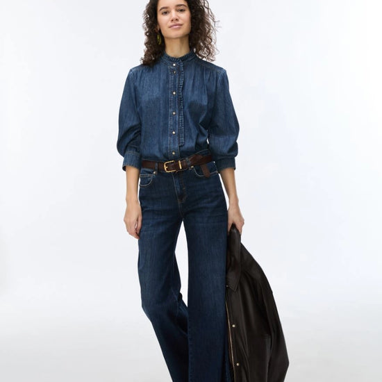 A model wearing a dark blue denim button-up shirt, matching wide-leg jeans, a brown belt, and holding a black leather jacket against a plain white studio background.