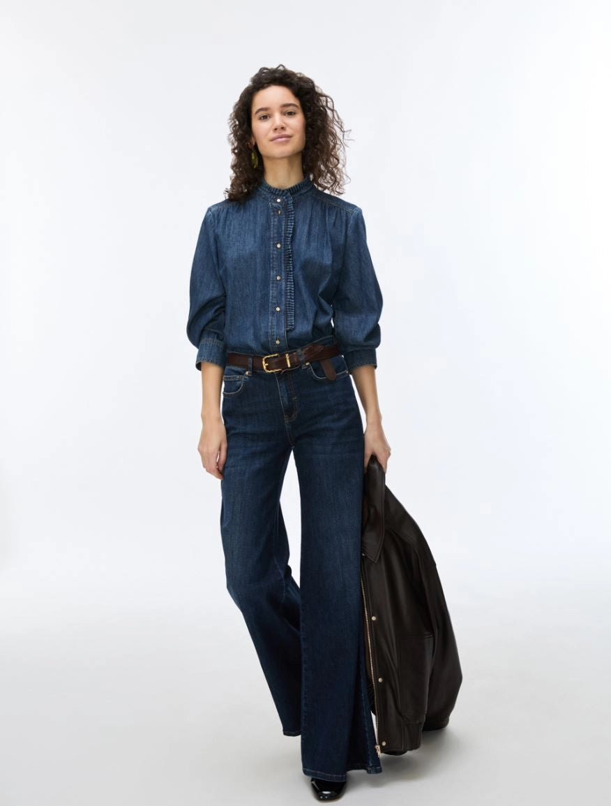 A model wearing a dark blue denim button-up shirt, matching wide-leg jeans, a brown belt, and holding a black leather jacket against a plain white studio background.