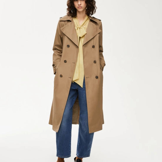 Person wearing a beige trench coat over a yellow shirt and blue jeans on a white background