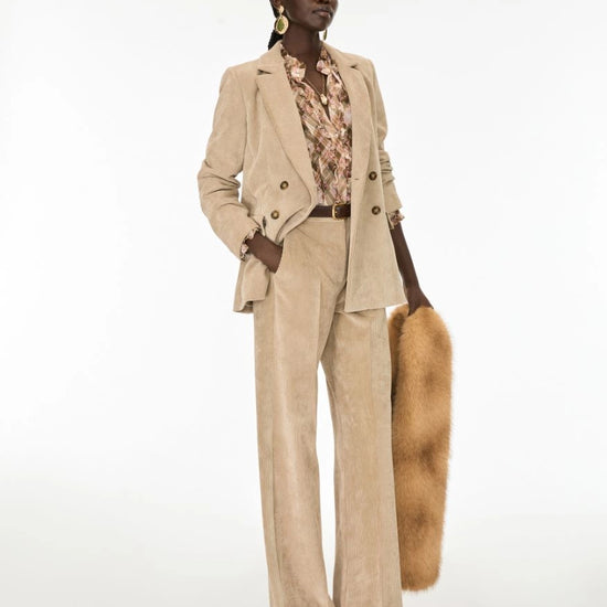 A model stands against a plain white studio background wearing a beige corduroy blazer and matching wide-leg trousers, layered over a floral shirt, holding a tan fur accessory.