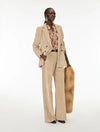 A model stands against a plain white studio background wearing a beige corduroy blazer and matching wide-leg trousers, layered over a floral shirt, holding a tan fur accessory.