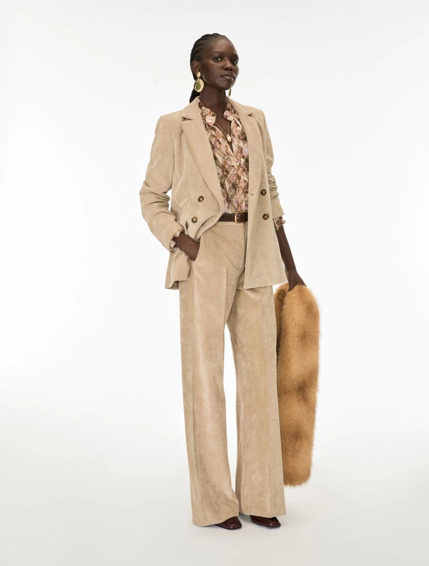 A model stands against a plain white studio background wearing a beige corduroy blazer and matching wide-leg trousers, layered over a floral shirt, holding a tan fur accessory.