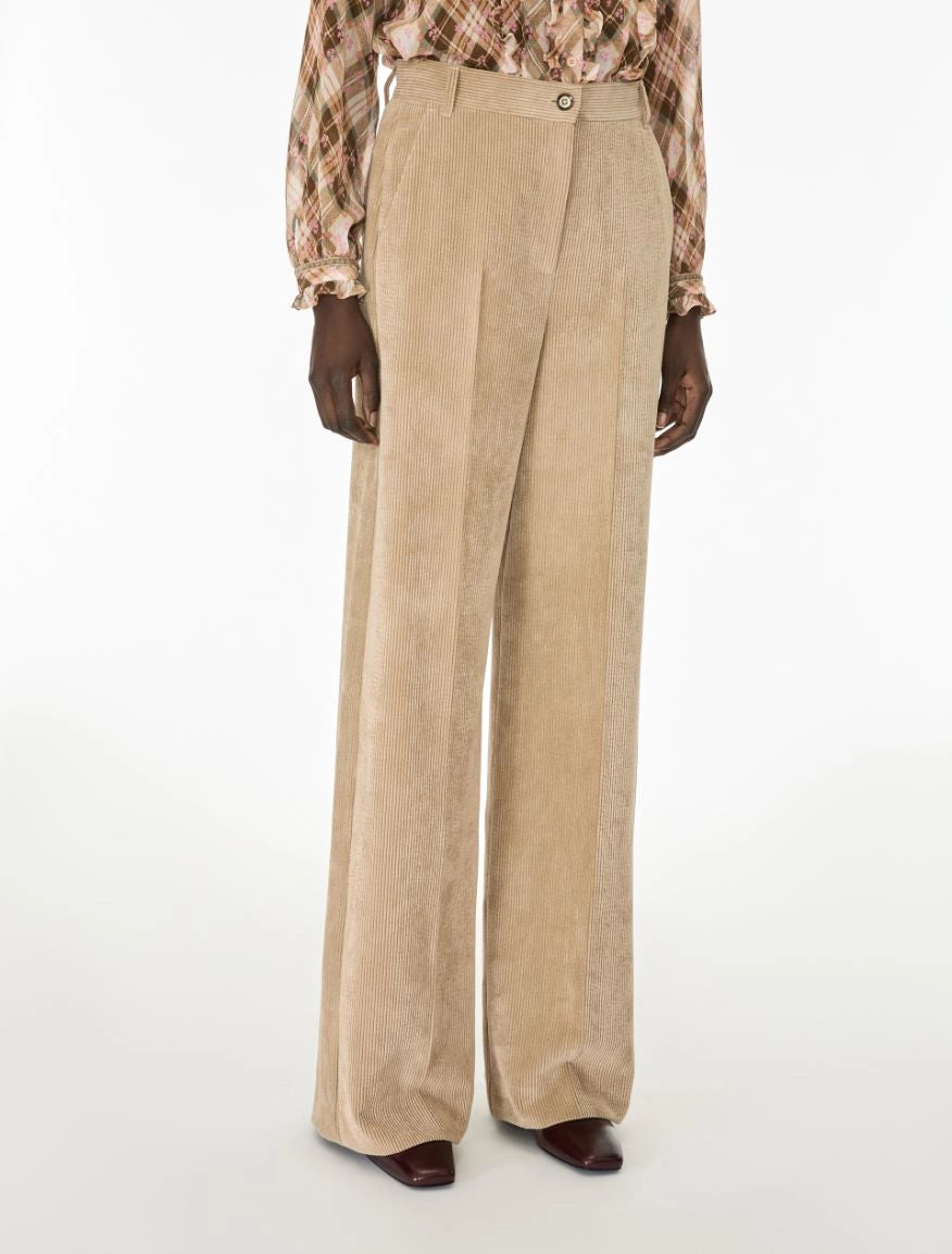 A model is shown from the waist down wearing high-waisted, wide-leg beige corduroy trousers, a brown and beige plaid shirt, and dark brown shoes against a plain white studio background.