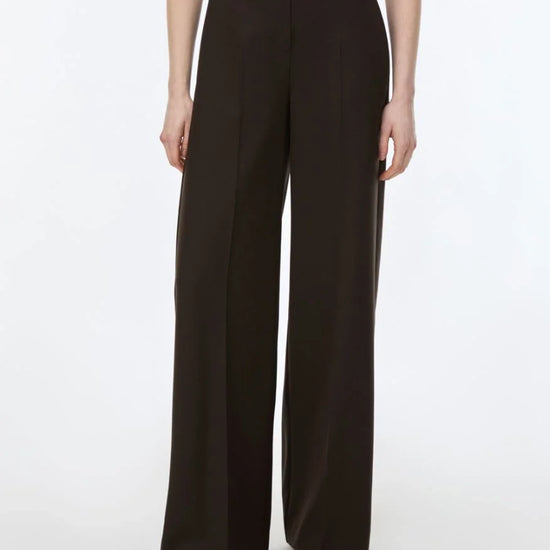 A model wearing high-waisted, wide-leg dark brown trousers, paired with a dark brown top and brown flat shoes, photographed against a clean white studio background.