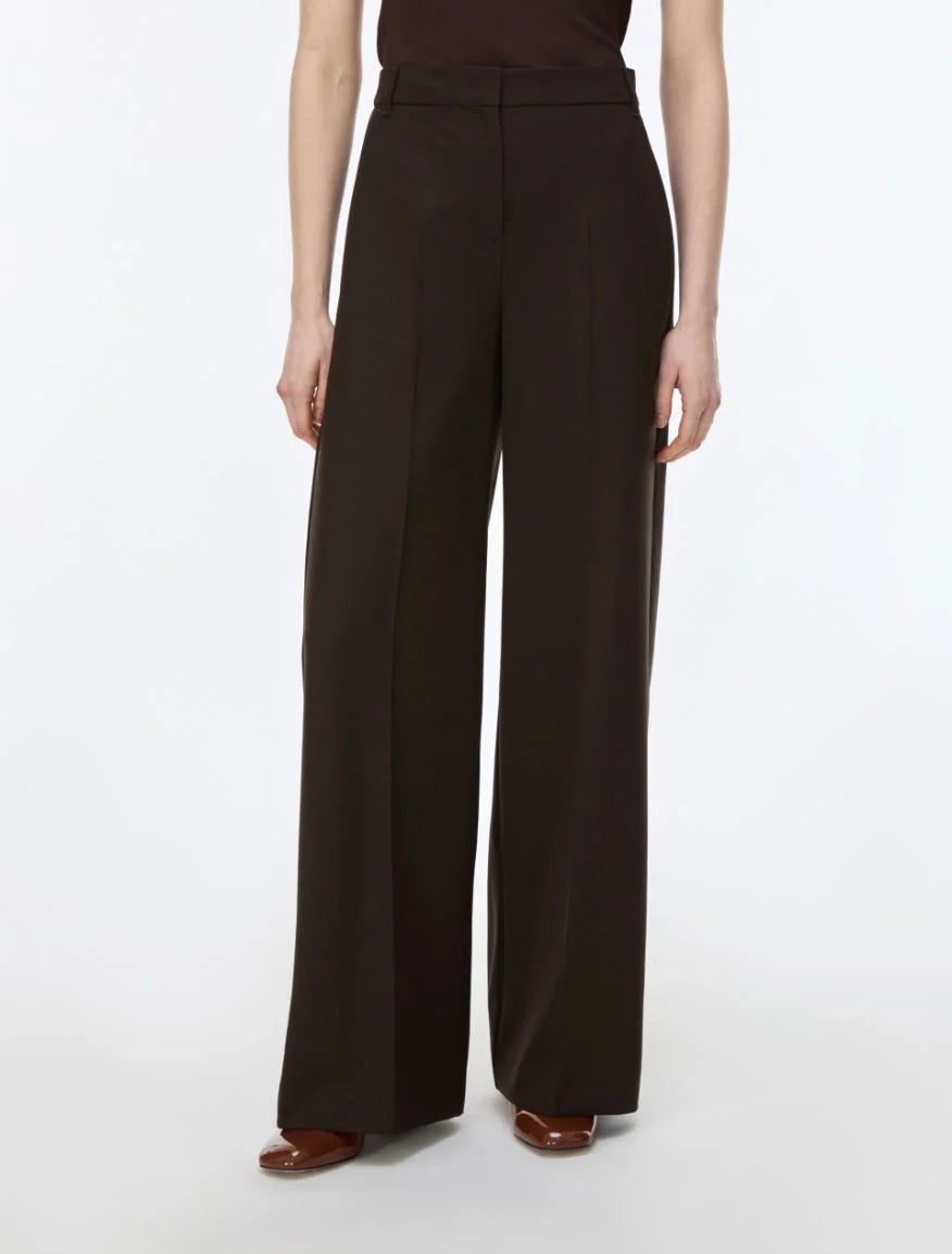 A model wearing high-waisted, wide-leg dark brown trousers, paired with a dark brown top and brown flat shoes, photographed against a clean white studio background.