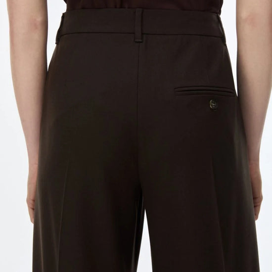 Close-up back view of a person wearing dark brown tailored trousers with a welt pocket and a small metallic button detail, paired with a dark top against a light gray background.