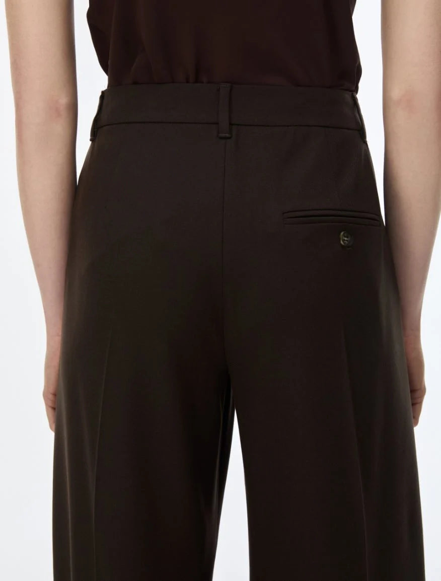 Close-up back view of a person wearing dark brown tailored trousers with a welt pocket and a small metallic button detail, paired with a dark top against a light gray background.