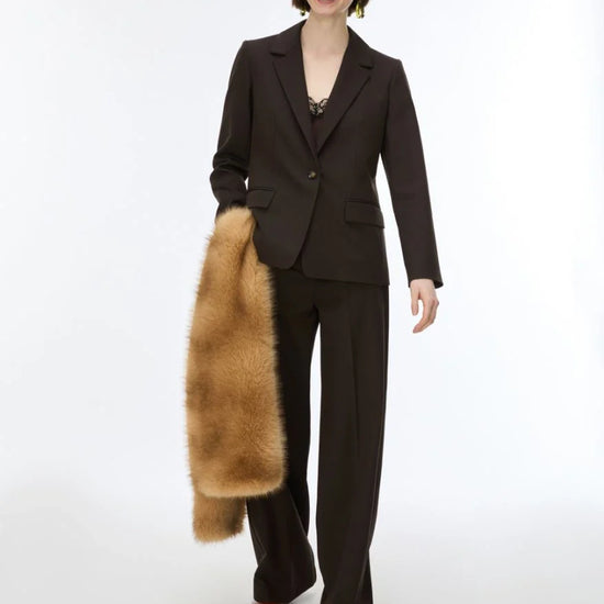 A model wearing a dark brown tailored suit with wide-leg trousers and a dark blazer, holding a tan fur coat against a plain white studio background.