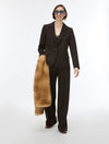 A model wearing a dark brown tailored suit with wide-leg trousers and a dark blazer, holding a tan fur coat against a plain white studio background.