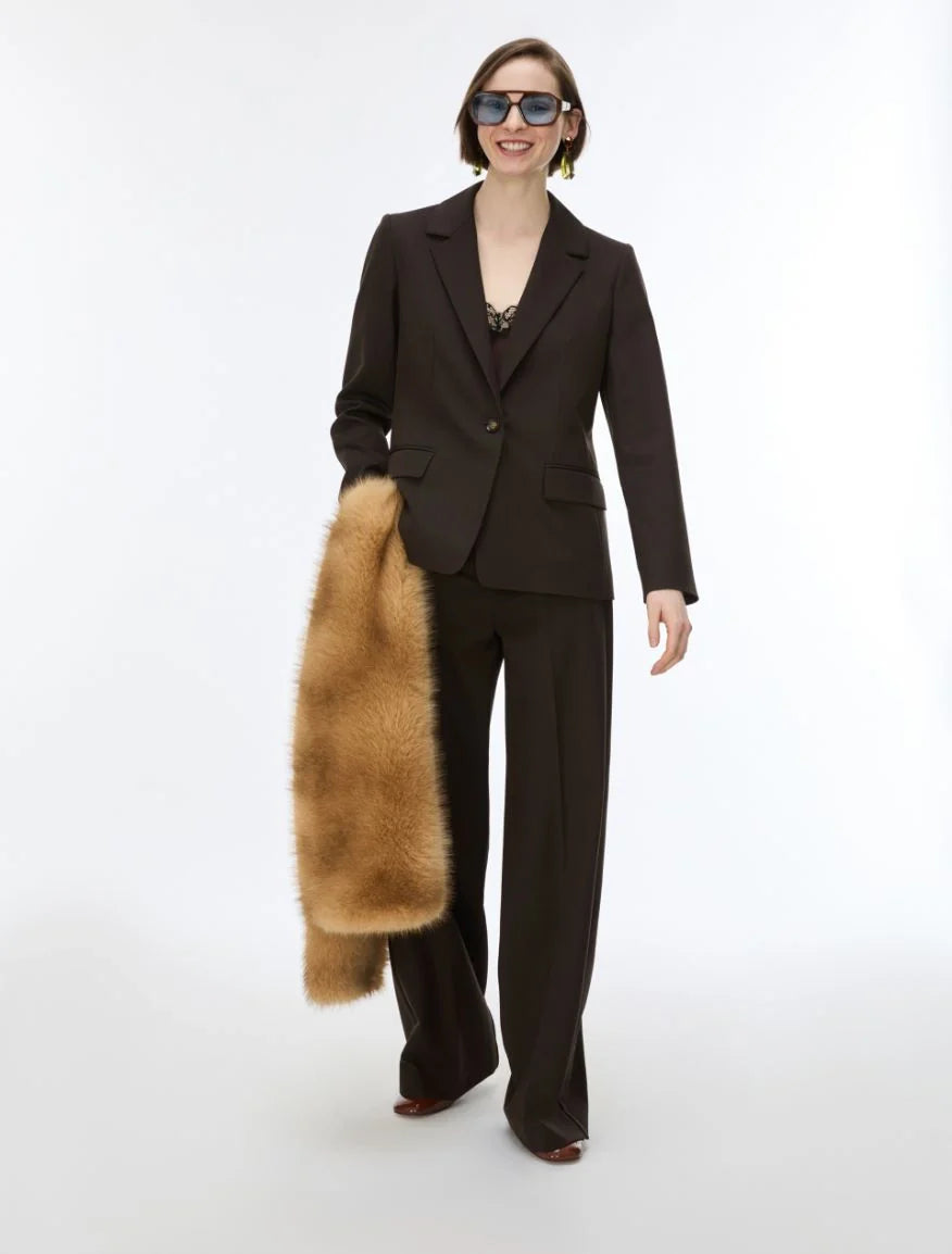 A model wearing a dark brown tailored suit with wide-leg trousers and a dark blazer, holding a tan fur coat against a plain white studio background.