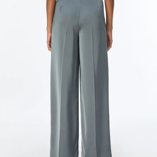 A model shown from the back wearing high-waisted, wide-leg trousers in a muted slate gray color, paired with a matching gray top and metallic heels, against a plain white studio background.