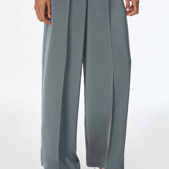 A person wearing high-waisted, wide-leg gray trousers with a smooth satin-like finish, paired with metallic silver heeled sandals against a plain white studio background.