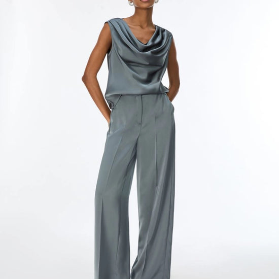 A woman modeling a slate blue-gray cowl-neck sleeveless jumpsuit with wide legs, paired with silver heeled sandals, against a plain white studio background.