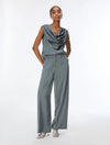 A woman modeling a slate blue-gray cowl-neck sleeveless jumpsuit with wide legs, paired with silver heeled sandals, against a plain white studio background.