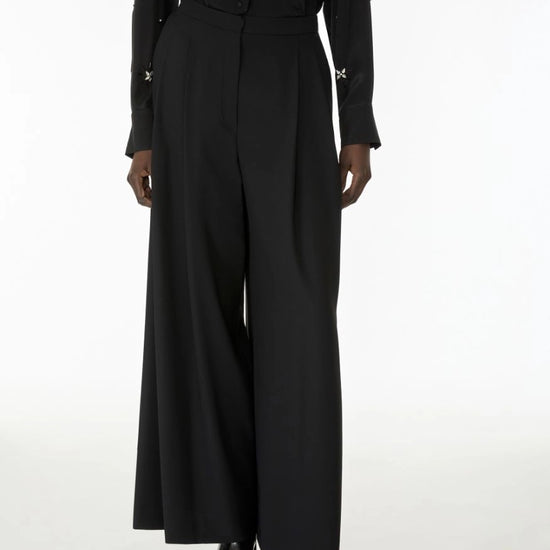 A model wearing a black long-sleeve button-front top with small metallic star details on the cuffs and waist, paired with high-waisted black wide-leg trousers and black shoes, against a plain white studio background.