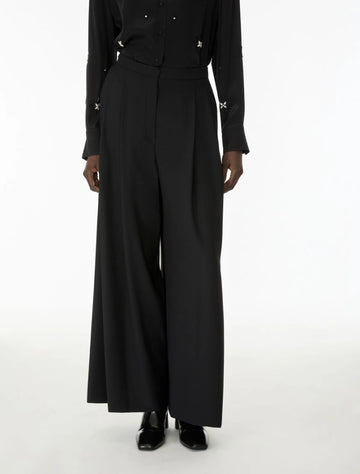 A model wearing a black long-sleeve button-front top with small metallic star details on the cuffs and waist, paired with high-waisted black wide-leg trousers and black shoes, against a plain white studio background.