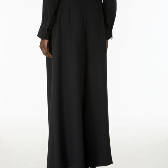 A person shown from the back wearing a long black dress with a high waist, long sleeves, and black heeled shoes against a plain white studio background.