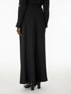 A person shown from the back wearing a long black dress with a high waist, long sleeves, and black heeled shoes against a plain white studio background.