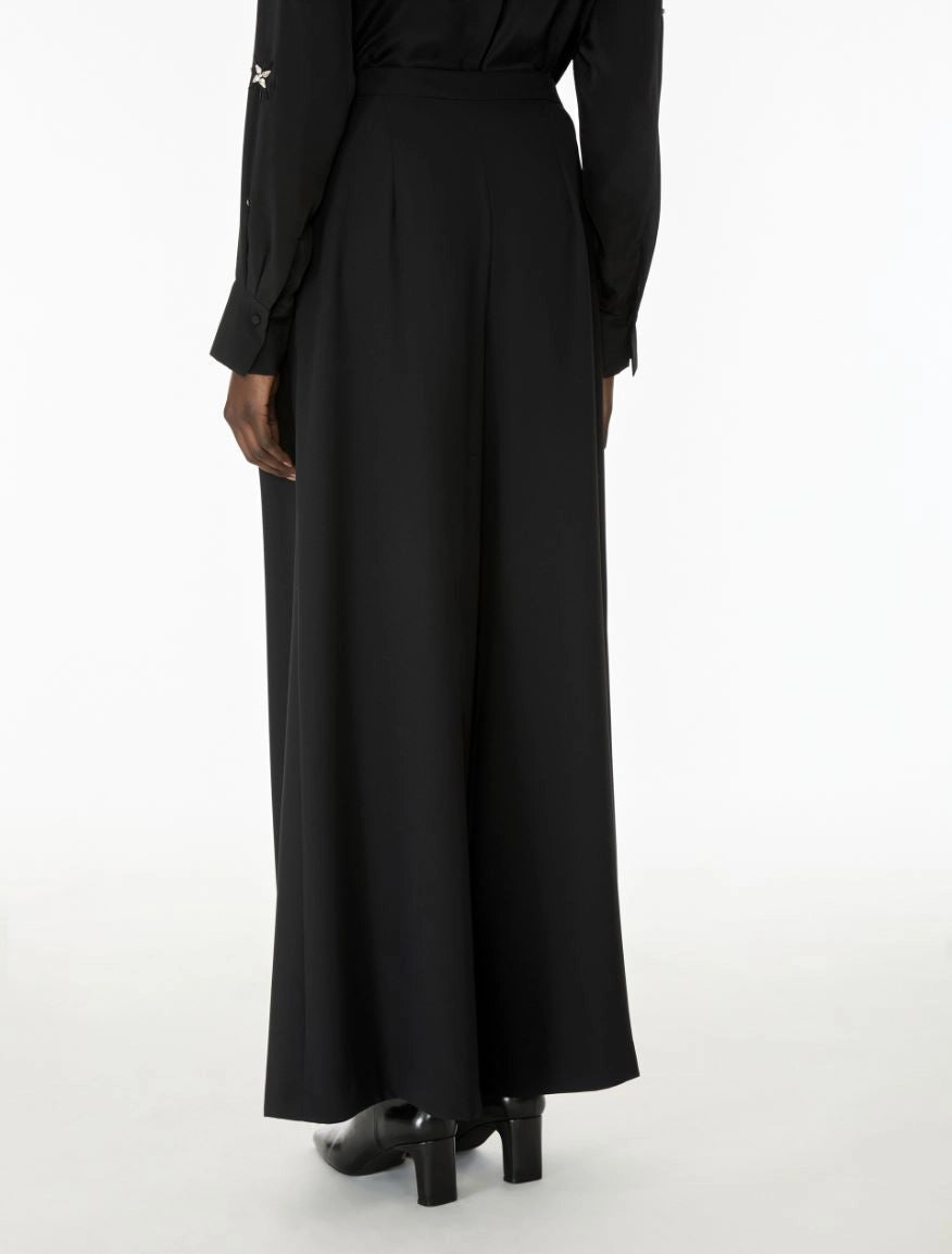A person shown from the back wearing a long black dress with a high waist, long sleeves, and black heeled shoes against a plain white studio background.