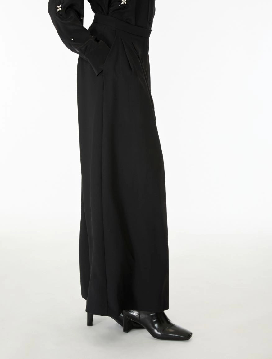 A model wearing a long black wide-leg dress with star details on the sleeves, paired with black pointed-toe heels, shown from the waist down against a plain white studio background.