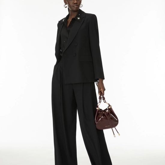 A model stands against a white studio background wearing a black double-breasted blazer and matching wide-leg trousers, holding a small dark brown leather bucket bag.