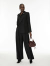 A model stands against a white studio background wearing a black double-breasted blazer and matching wide-leg trousers, holding a small dark brown leather bucket bag.