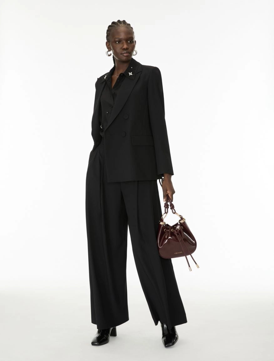 A model stands against a white studio background wearing a black double-breasted blazer and matching wide-leg trousers, holding a small dark brown leather bucket bag.