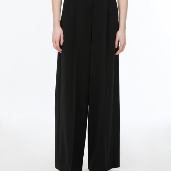 A model wearing high-waisted, wide-leg black trousers with a flat front and side pockets, paired with a beige top and metallic sandals, against a plain white studio background.