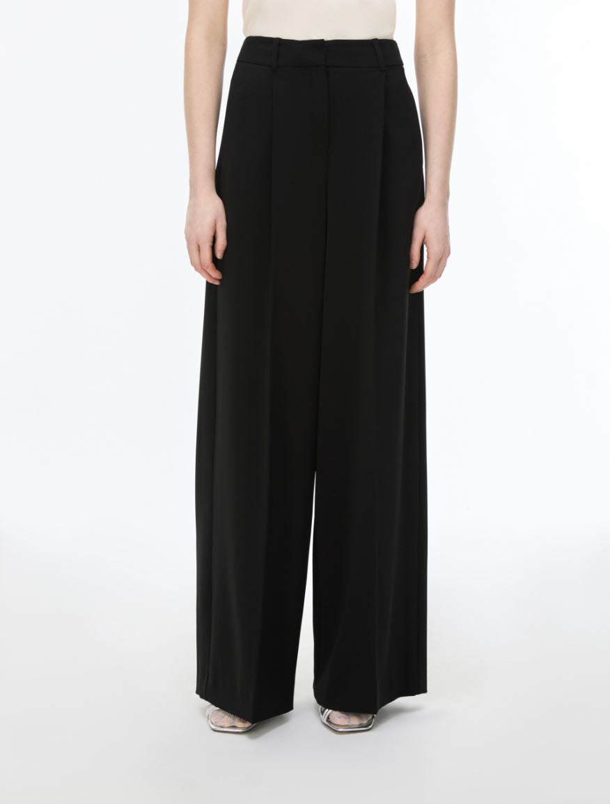 A model wearing high-waisted, wide-leg black trousers with a flat front and side pockets, paired with a beige top and metallic sandals, against a plain white studio background.