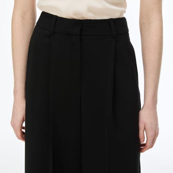 A close-up of a model wearing black wide-leg pleated trousers with a matching black belt, paired with a cream-colored satin blouse, against a plain white background.