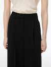A close-up of a model wearing black wide-leg pleated trousers with a matching black belt, paired with a cream-colored satin blouse, against a plain white background.