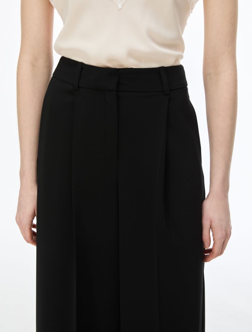 A close-up of a model wearing black wide-leg pleated trousers with a matching black belt, paired with a cream-colored satin blouse, against a plain white background.