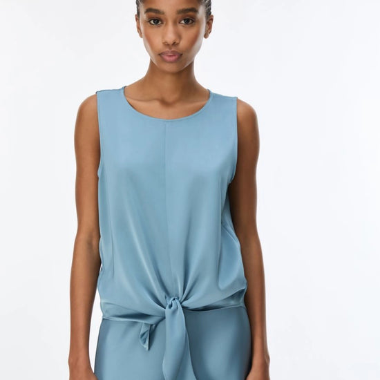 A woman modeling a sleeveless light blue top with a gathered waist and front tie detail, photographed against a plain white studio background.