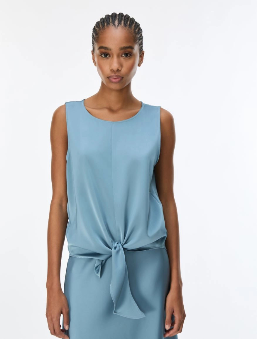A woman modeling a sleeveless light blue top with a gathered waist and front tie detail, photographed against a plain white studio background.