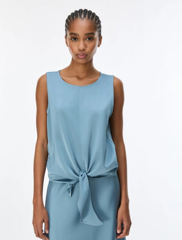 A woman modeling a sleeveless light blue top with a gathered waist and front tie detail, photographed against a plain white studio background.
