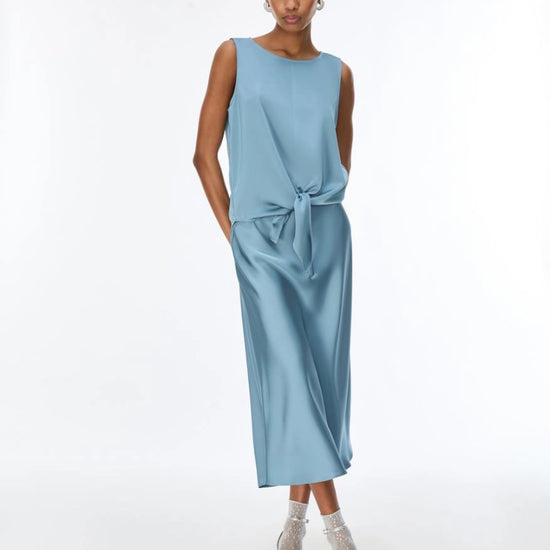 A woman modeling a sleeveless light blue top with a tied waist, paired with clear high-heeled sandals, against a plain white studio background.