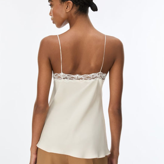 A woman shown from the back wearing a cream-colored satin camisole top with thin straps and a lace trim across the upper back, paired with tan pants, against a plain white background.