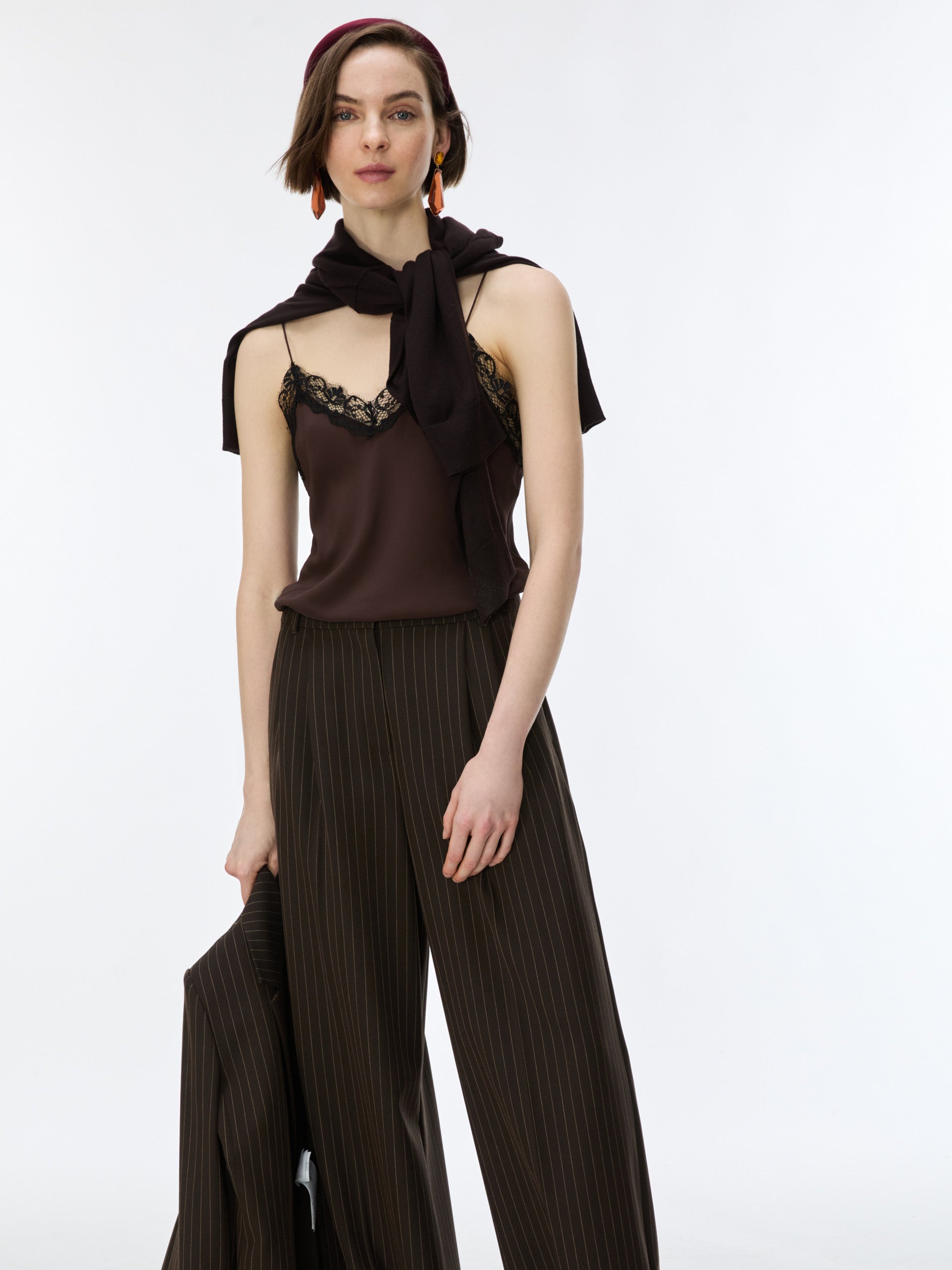 A model wearing a dark brown sleeveless top with lace trim and a matching draped scarf, paired with dark brown pleated wide-leg trousers, against a plain white studio background.