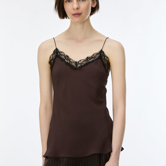 A woman modeling a dark brown satin camisole top with black lace trim along the neckline, paired with dark pinstriped trousers, against a plain white studio background.