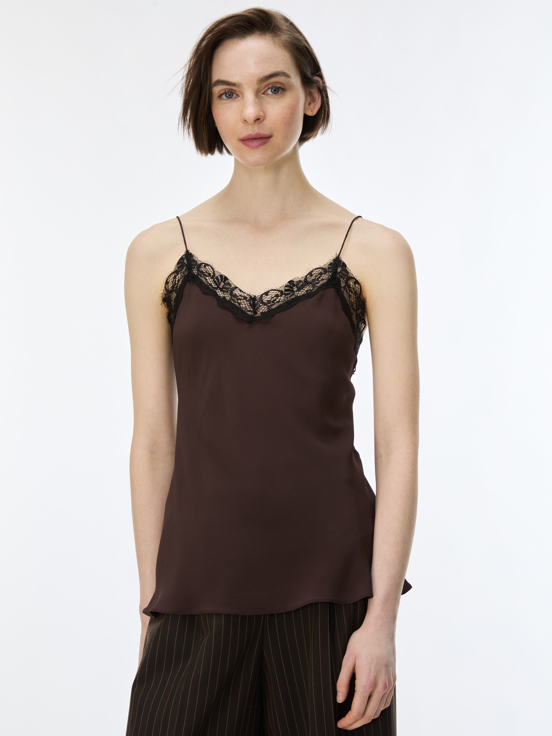 A woman modeling a dark brown satin camisole top with black lace trim along the neckline, paired with dark pinstriped trousers, against a plain white studio background.