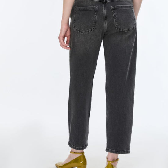 Rear view of a person wearing dark gray washed cropped jeans with a small tan leather patch on the back waistband, paired with metallic gold heels against a plain white studio background.