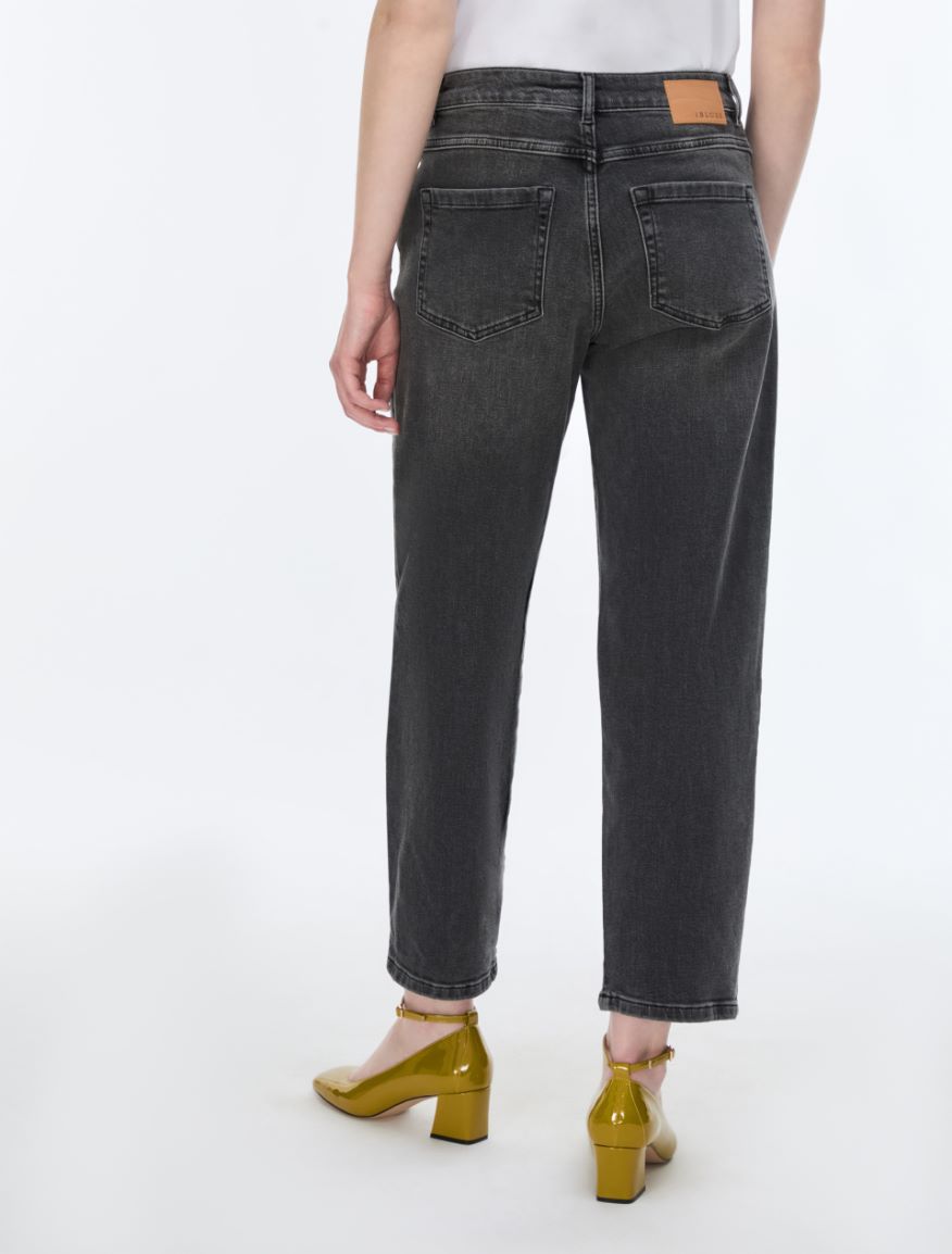 Rear view of a person wearing dark gray washed cropped jeans with a small tan leather patch on the back waistband, paired with metallic gold heels against a plain white studio background.