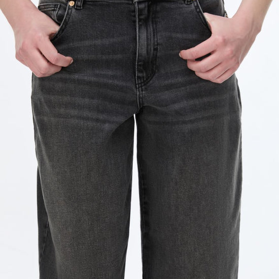 A close-up of a person wearing dark gray washed denim jeans with hands in the pockets, against a plain white background.
