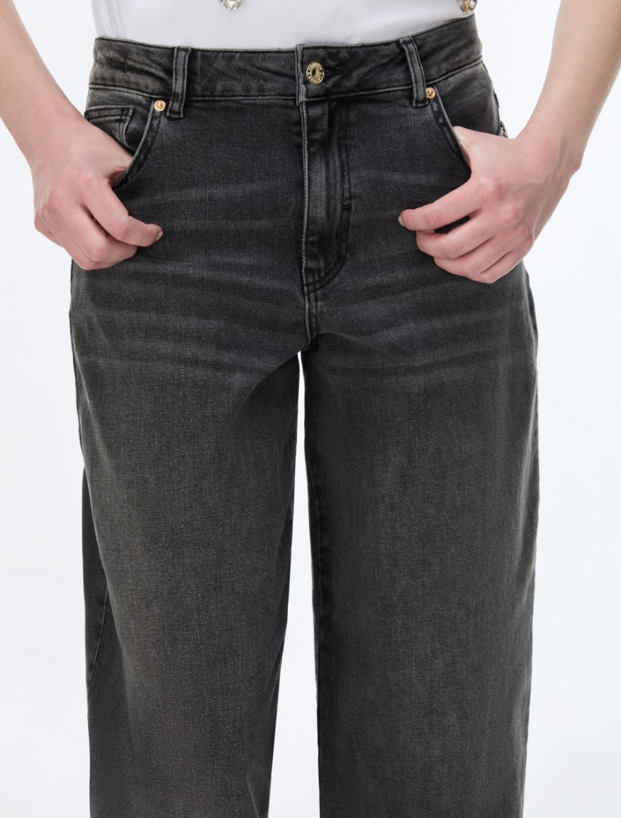A close-up of a person wearing dark gray washed denim jeans with hands in the pockets, against a plain white background.
